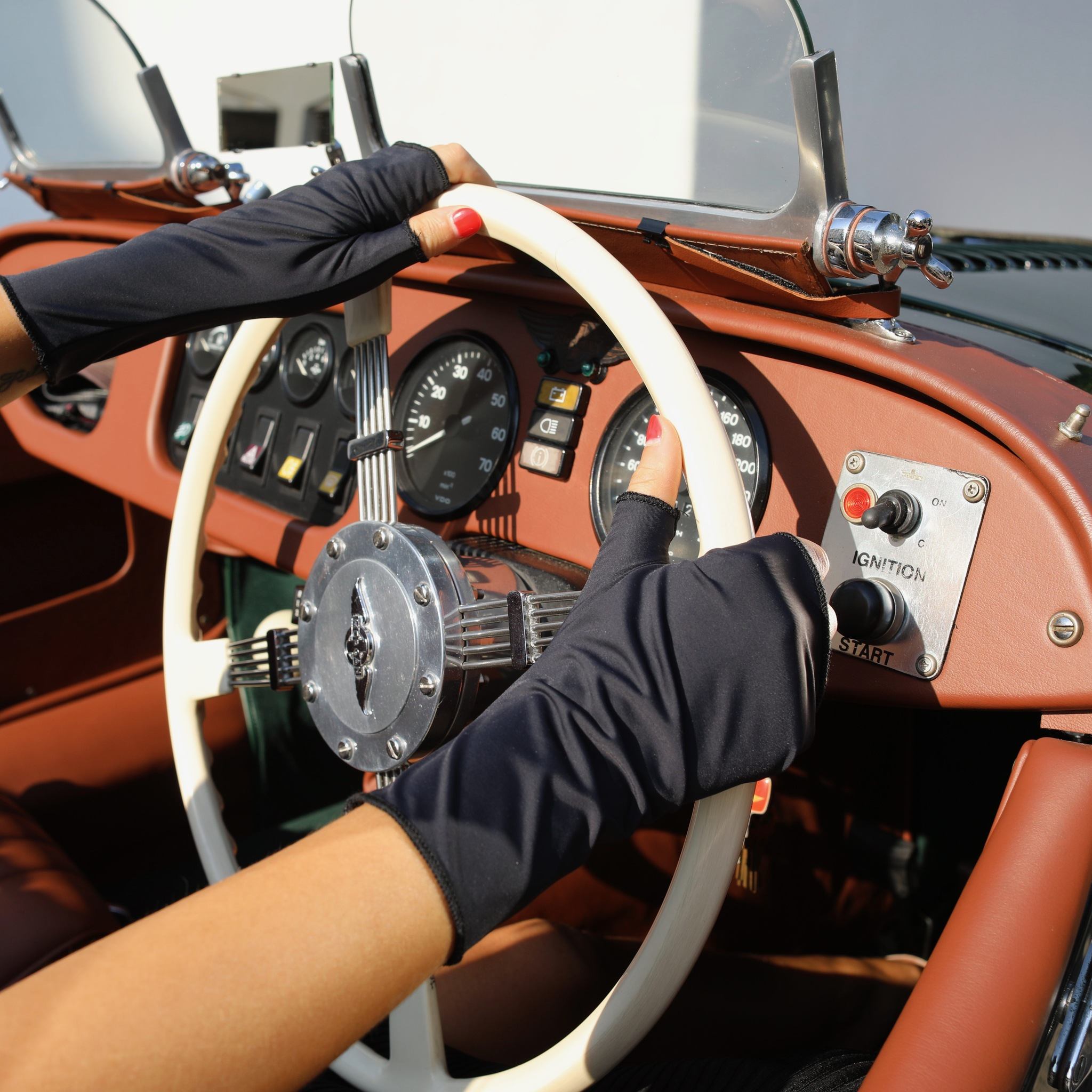 Woman holding a car steering wheel wearing short black UPF 50+ (sun protective) gloves.