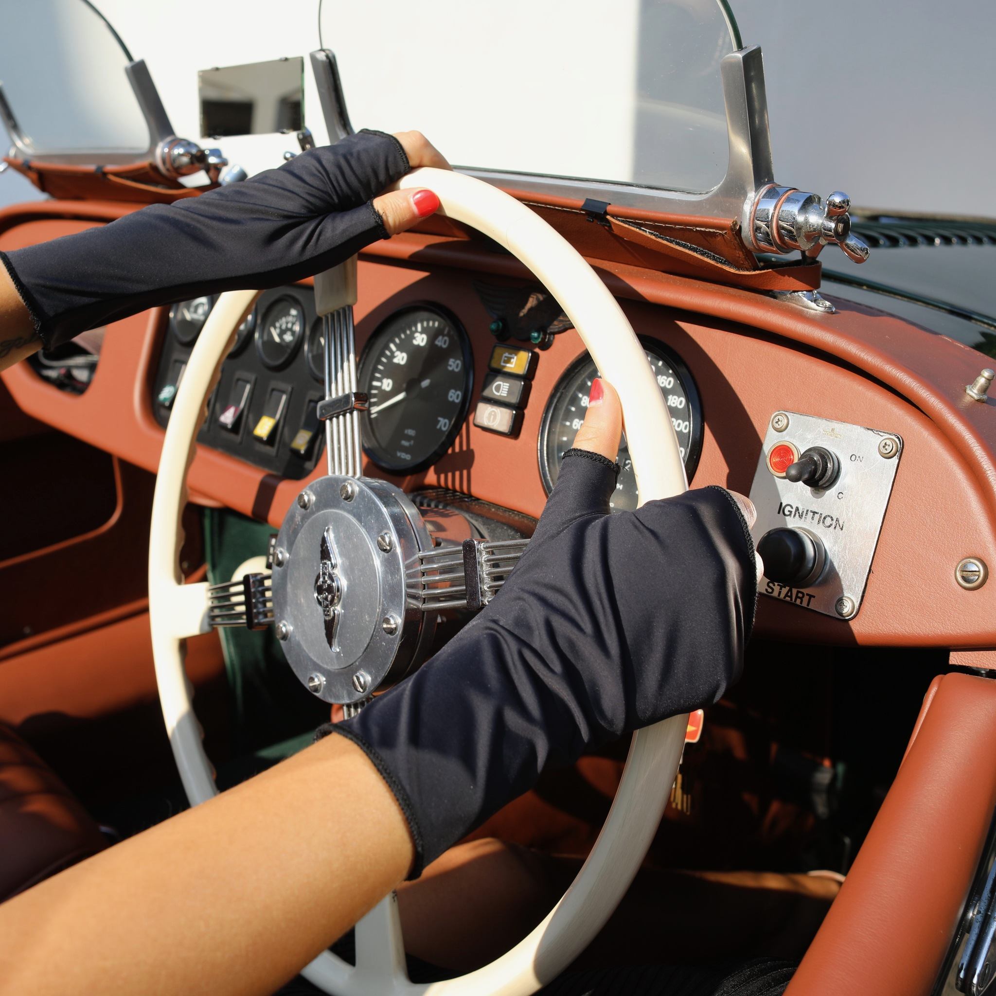 Woman holding a car steering wheel wearing short black UPF 50+ (sun protective) gloves. 