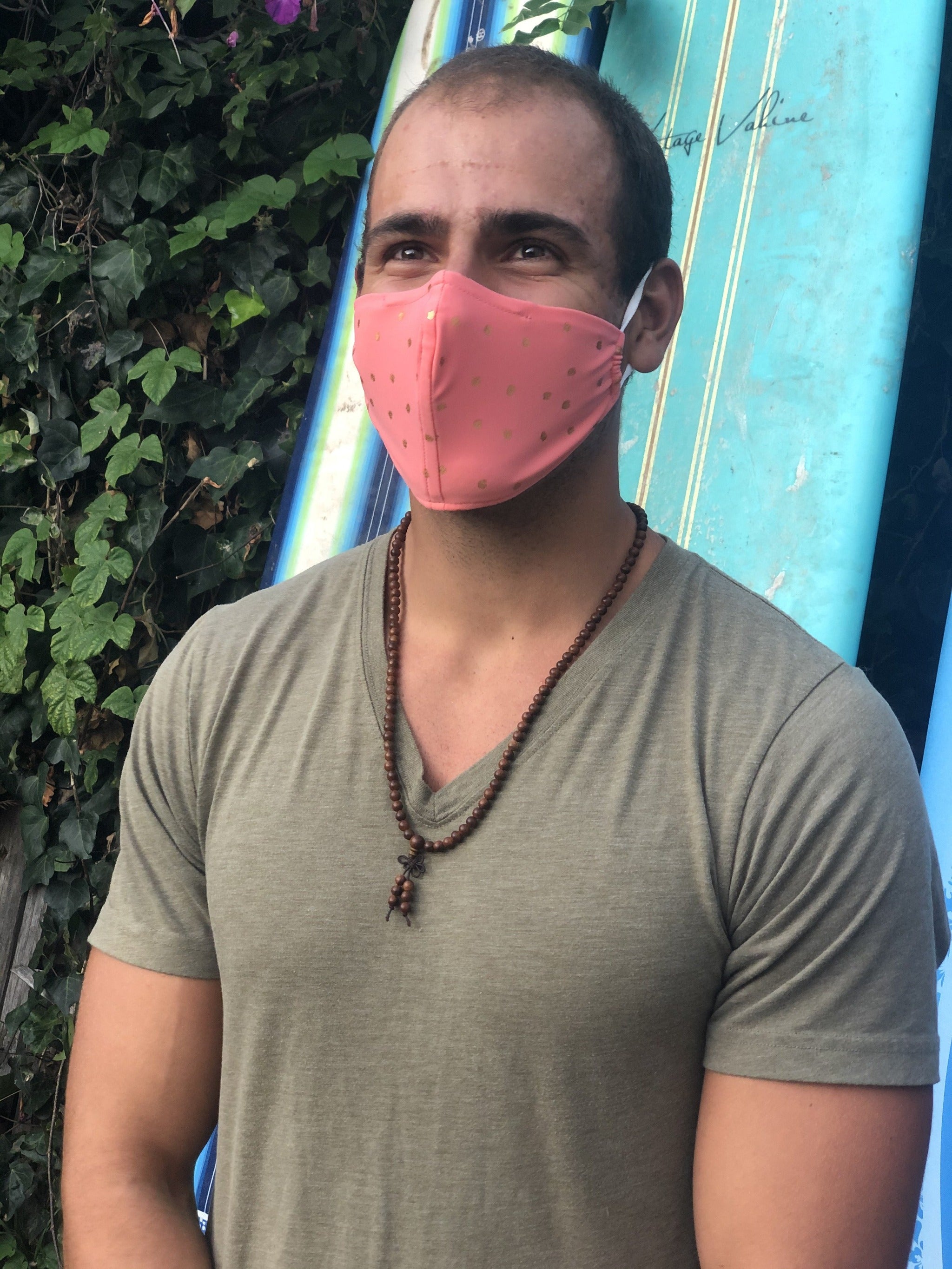 Man in front of surfboards wearing a pink face mask with gold polka dots.