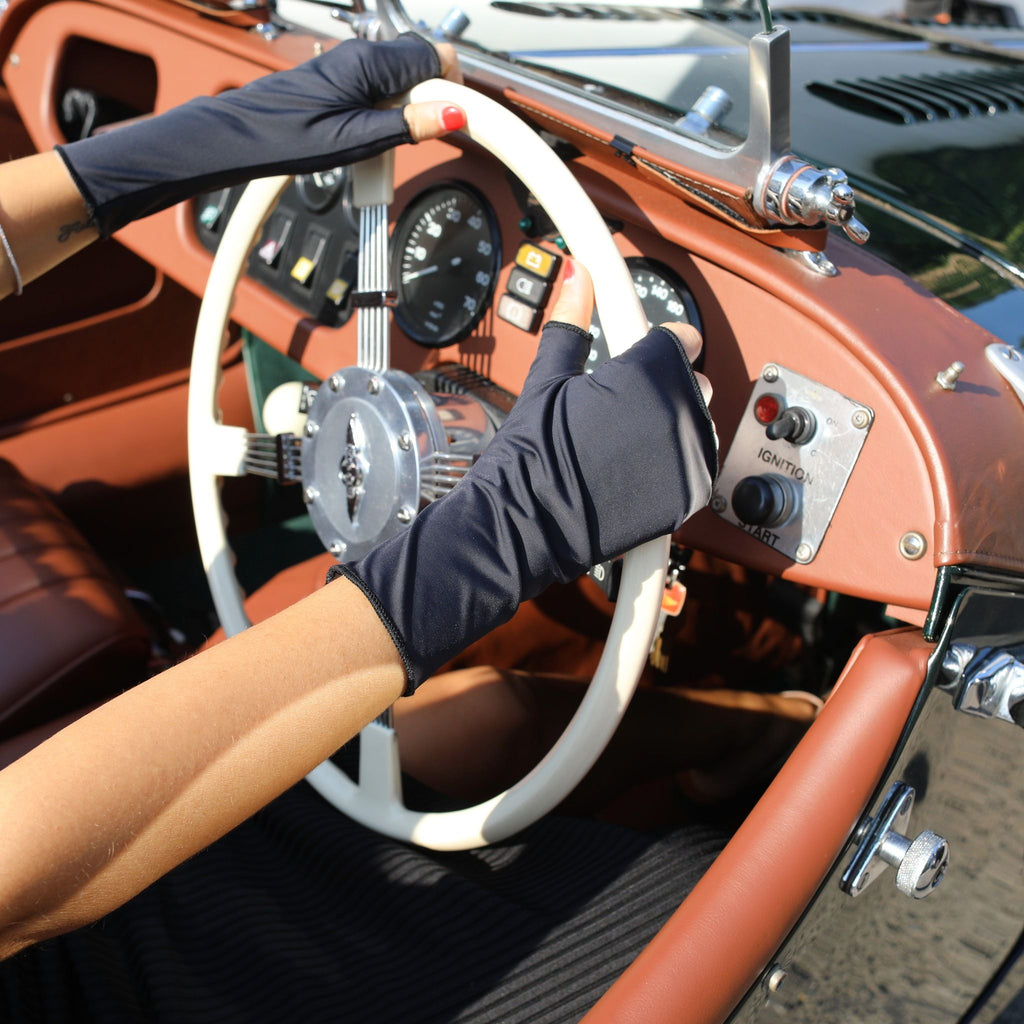 Woman holding a car steering wheel wearing short black UPF 50+ (sun protective) gloves. 