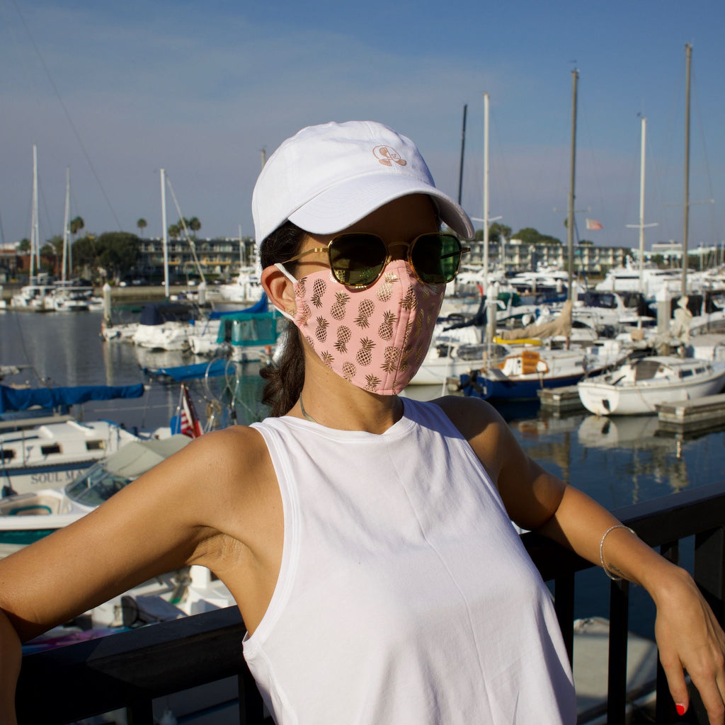 Woman in a sunny boat-filled marina wearing a pink face mask with gold pineapples.