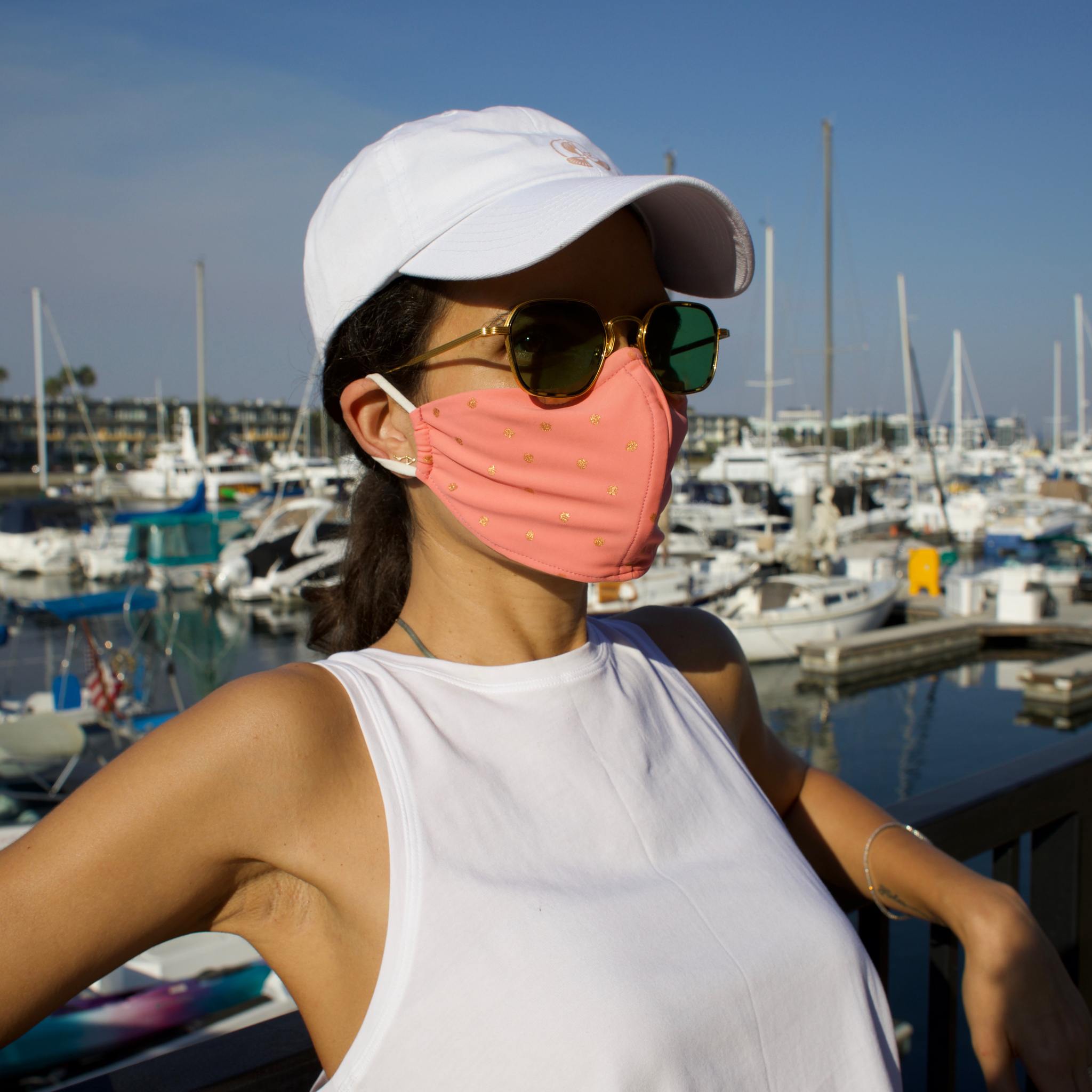 Woman in a sunny boat-filled marina wearing a pink face mask with gold polka dots.