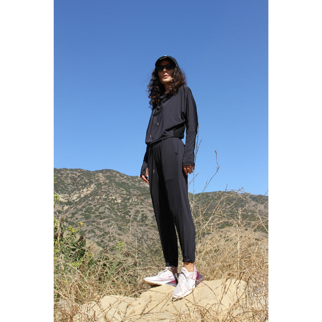 Woman hiking, wearing a lightweight long sleeved sun protective hoodie with hand coverings and matching UPF 50+ drawstring pants in black.