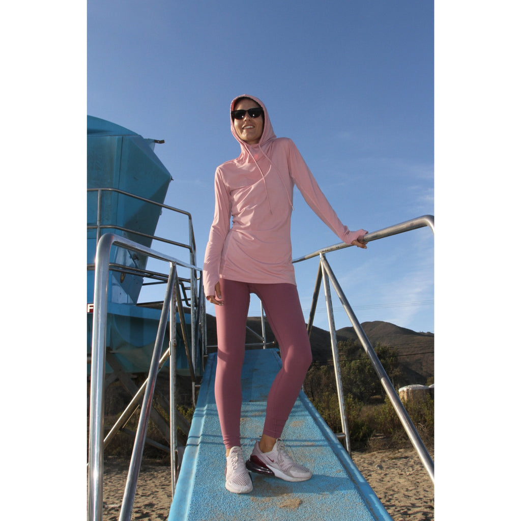 Woman on a beach wearing a long-sleeved UPF 50+ (sun protective) shirt with full hood and hand protection in pink/blush.