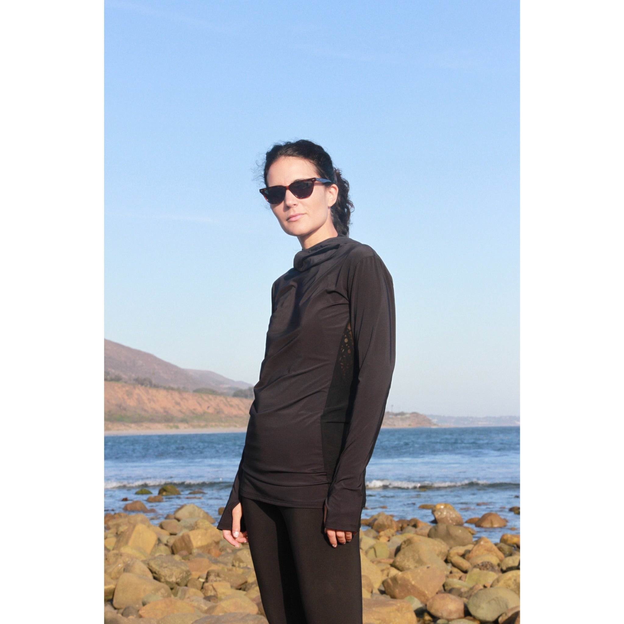 Woman on a beach wearing a long-sleeved UPF 50+ (sun protective) shirt with full hood and hand protection in black.