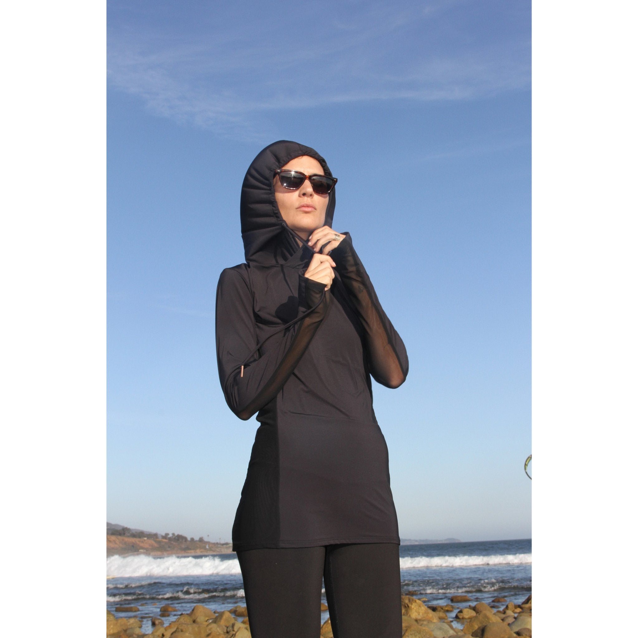 Woman on a beach wearing a long-sleeved UPF 50+ (sun protective) shirt with full hood and hand protection in black.