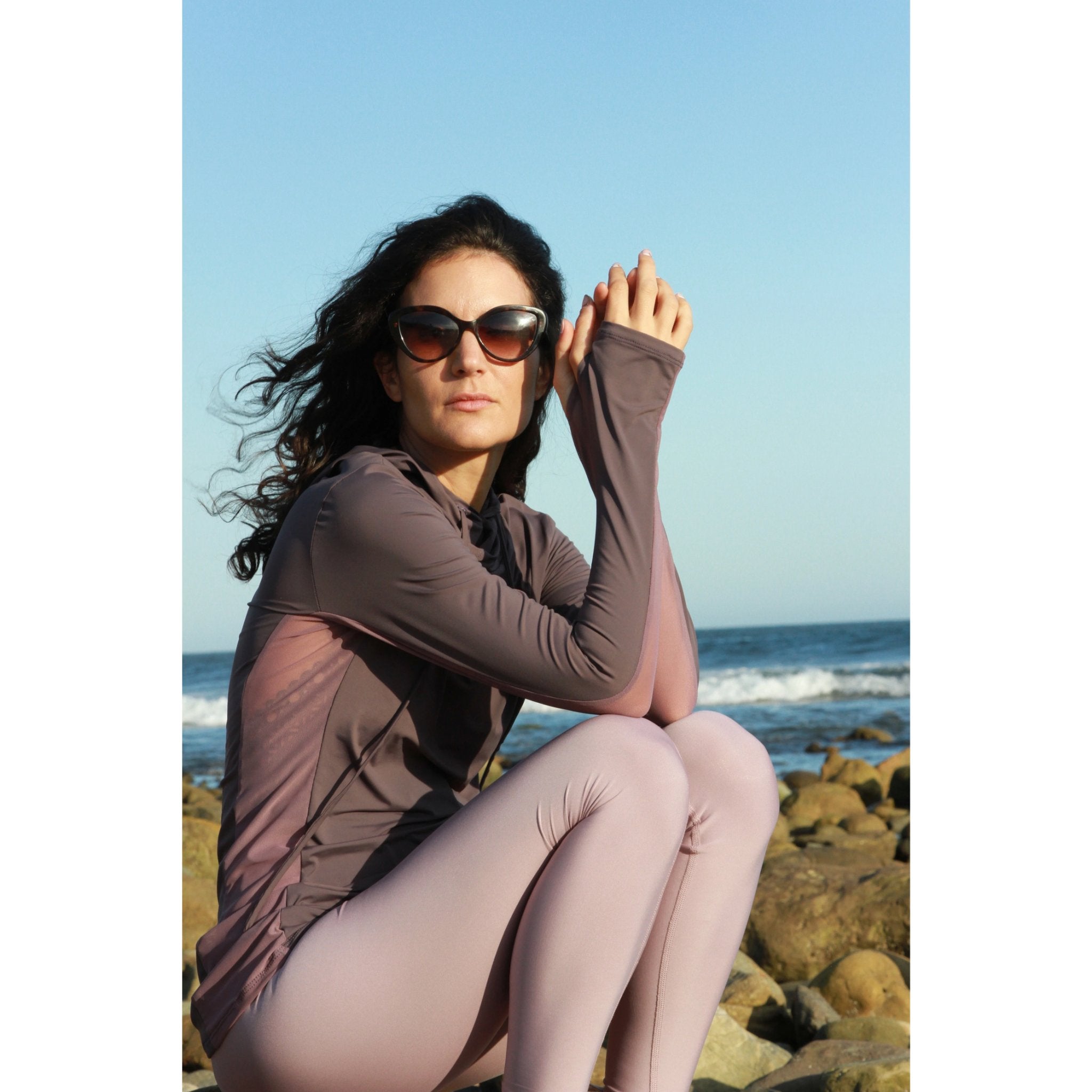 Woman on a beach wearing a long-sleeved UPF 50+ (sun protective) shirt with full hood and hand protection in plum.