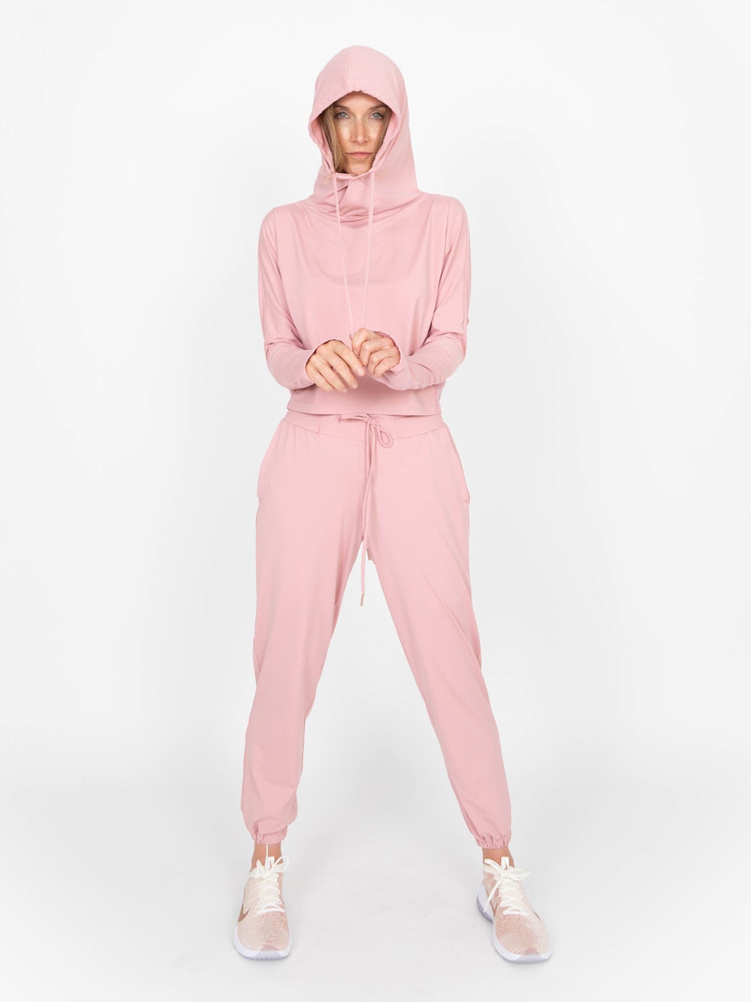 Woman wearing a lightweight long sleeved sun protective hoodie with hand coverings in blush pink and matching drawstring pants. 